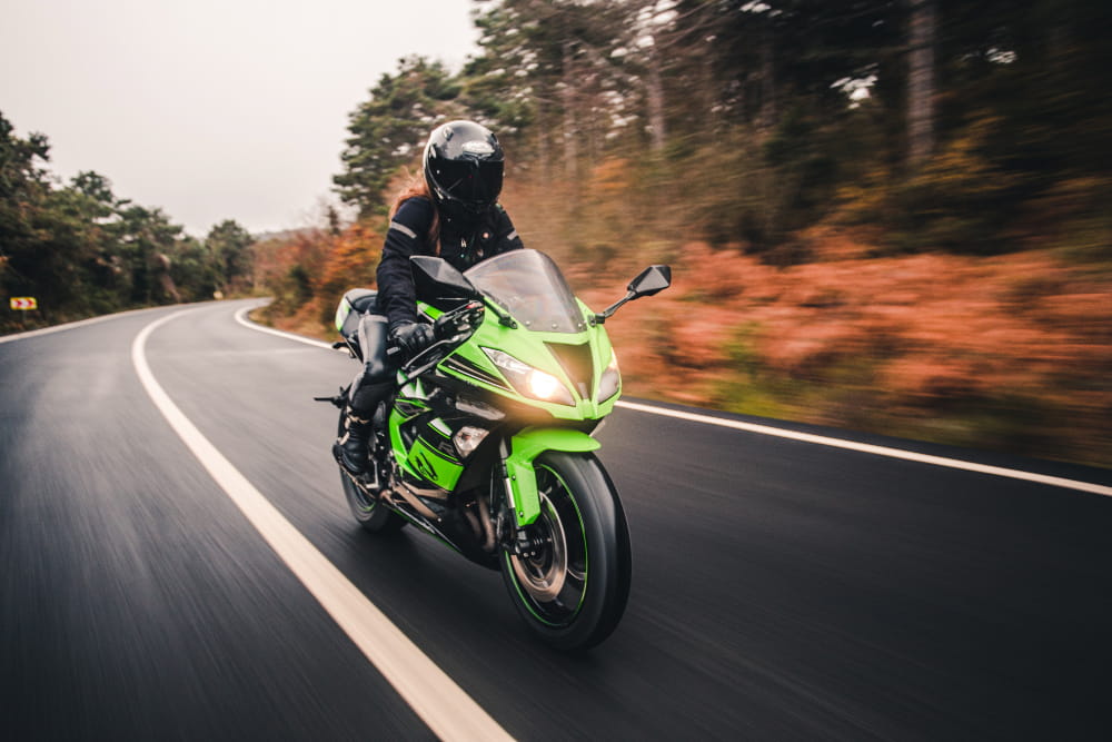 Motarde équipée d’un casque intégral conduisant une moto sportive verte sur une route sinueuse bordée d’arbres.